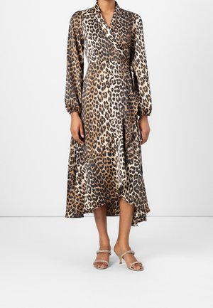 Frau trägt ein Wickelkleid mit langen Ärmeln in braun-schwarzem Leopardenmuster, kombiniert mit beigen offenen Sandalen mit Absatz, stehend auf weißem Boden.