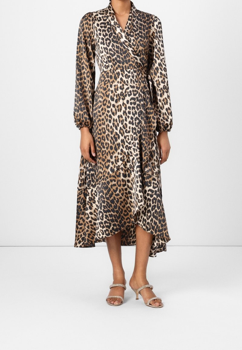 Femme portant une robe portefeuille à manches longues avec un imprimé léopard marron et noir, associée à des sandales à talons ouvertes beige, debout sur un sol blanc.