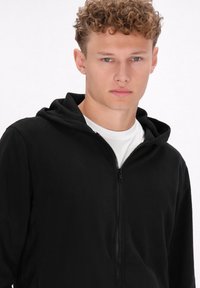 Črna zip-up kapuca iz mehkega flisa, ki ima kapuco in popoln sprednji zadrgo, oblečena čez belo majico. Enostaven in sproščen dizajn.