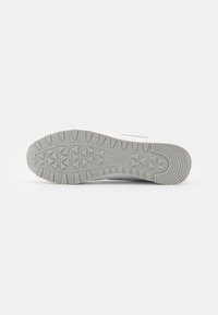 Hvit sneakersåle med en teksturert grå gummioverflate, med vinkelformede mønster for grep og en logo på siden.