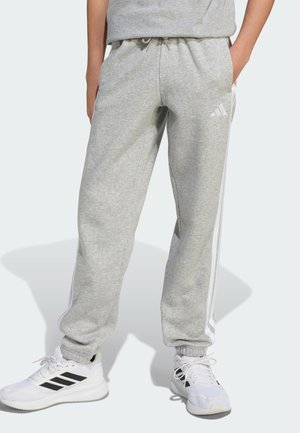 Persona con pantalones deportivos Adidas gris claro con rayas blancas laterales y zapatillas Adidas blancas con rayas negras, de pie con las manos en los bolsillos.