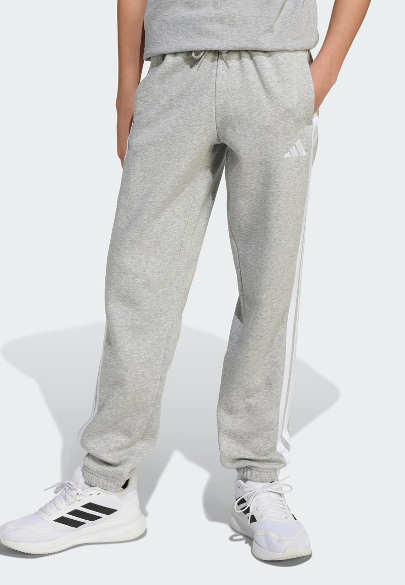Άτομο που φορά ανοιχτό γκρι φόρμα Adidas με λευκές πλευρικές ρίγες και λευκά sneakers Adidas με μαύρες ρίγες, στέκεται με τα χέρια στις τσέπες.
