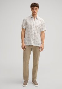 Kurzes, ärmelloses Hemd mit einem beigefarbenen Blumendruck, aus leichtem Stoff. Kombiniert mit khakifarbenen Hose und strukturierten Slippern.