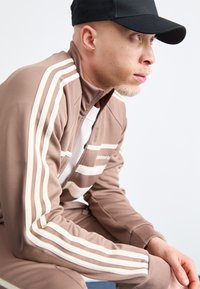 Brown tracksuit with white stripes, zip-up collar, long sleeves, and a relaxed fit. Black cap; smooth fabric texture.