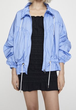 Person trägt eine hellblaue Reißverschlussjacke über einem schwarzen gerafften trägerlosen Kleid, gezeigt von den Schultern bis zur Mitte des Oberschenkels vor einfarbigem Hintergrund.