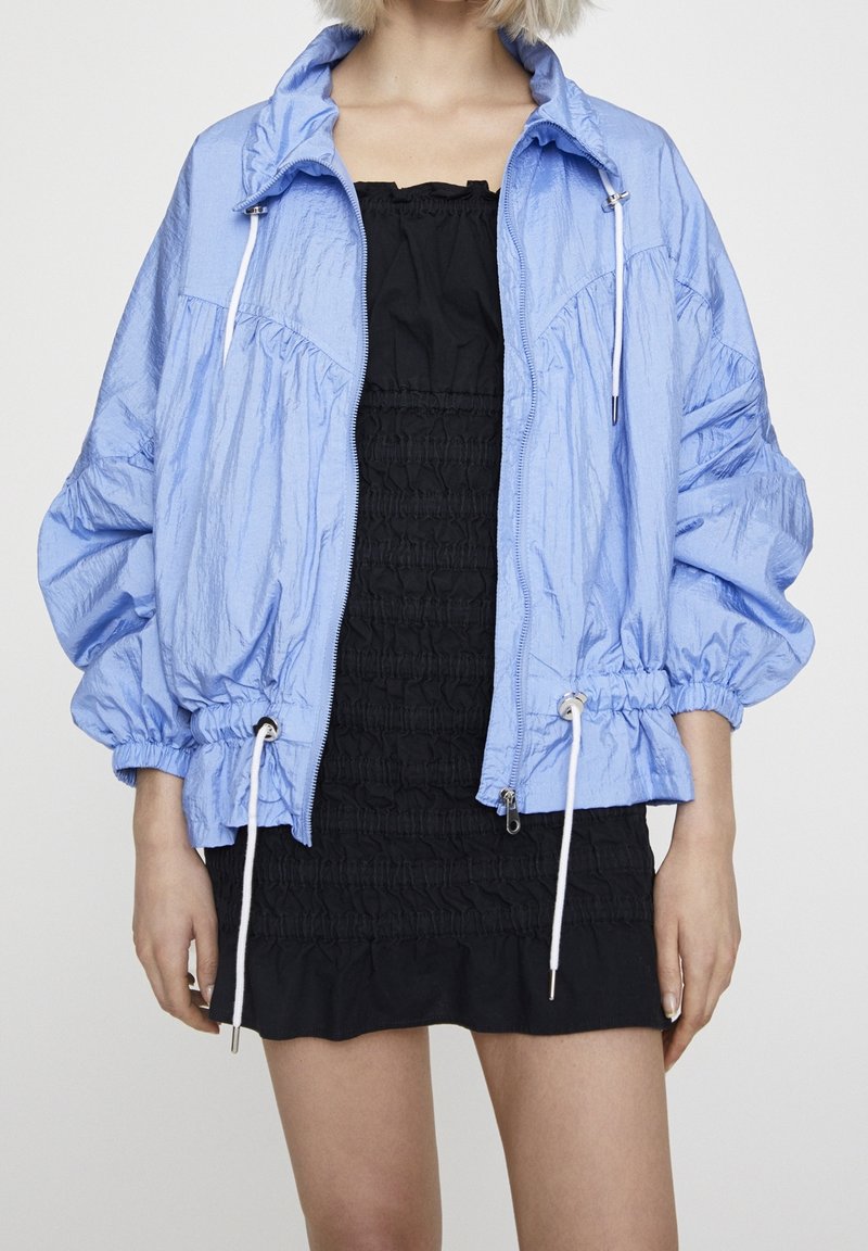 Personne portant une veste zippée bleu clair par-dessus une robe bustier noire froncée, visible des épaules jusqu'à mi-cuisse sur un fond uni.