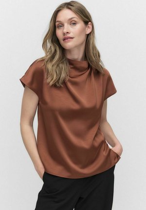 Brown satin short-sleeve top with a draped neckline and relaxed fit. Model stands with hands in pockets, wearing black pants. Smooth texture.