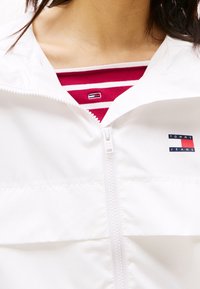 Witte jas met rits en een gestructureerde stof, met een klein blauw-rood "TOMMY JEANS" logo en een rood gestreept shirt eronder.