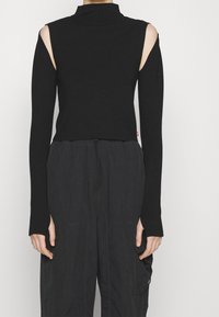 Schwarzes Langarmshirt, hoher Kragen, ausgeschnittene Schultern, figurbetontes Design, weiche Textur, getragen mit lockeren dunkelgrauen Hosen mit Kordelzug und Taschen.