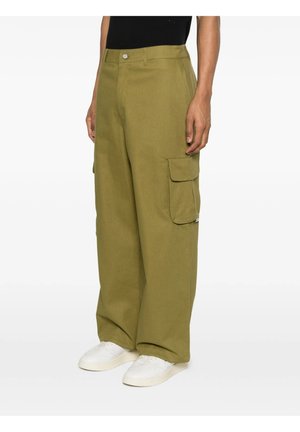 Uomo che indossa pantaloni cargo verde oliva a gamba larga con patta sulla tasca laterale e sneakers bianche, in piedi su uno sfondo semplice.