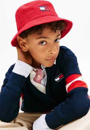 Fant, ki nosi rdeč teksturiran klobuk bucket in temno modro majico Tommy Hilfiger z rokavi v rdeče-bele-modri črti, gleda v kamero.
