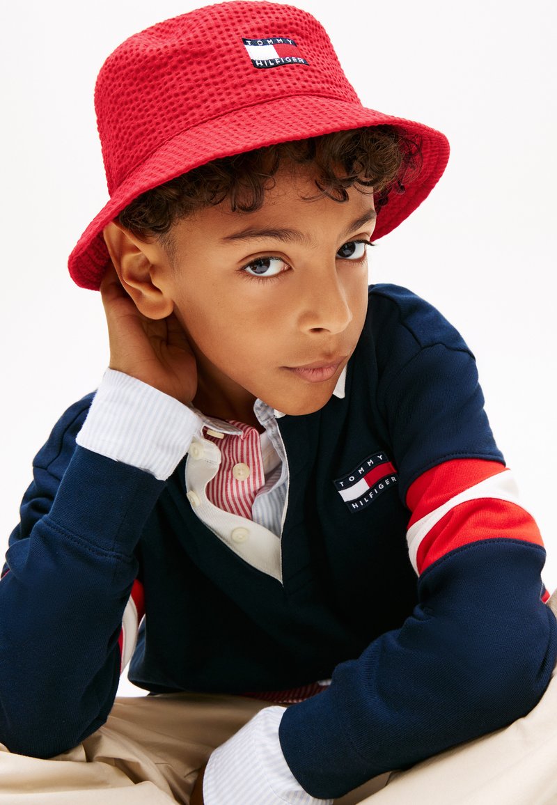 Αγόρι που φοράει κόκκινο υφασμάτινο καπέλο τύπου bucket και μπλε σκούρο πουκάμισο Tommy Hilfiger με μανίκια ριγέ κόκκινου, άσπρου και μπλε, κοιτάζοντας την κάμερα.