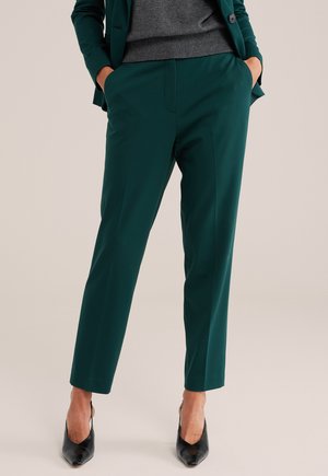Personne portant un pantalon vert foncé sur mesure, un pull gris et un blazer vert, les mains dans les poches et des chaussures noires à bout pointu.