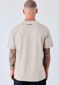 Camiseta de algodón beige de manga corta, con un ajuste regular y el logo negro "PXP" en la parte posterior. Textura suave y diseño sencillo.