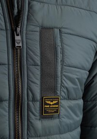 Giubbotto puffer di un intenso verde scuro con un distintivo patch intrecciato nero, logo dorato e una cerniera contrastante con un accento arancione.