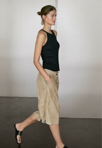 Černý tílko bez rukávů v kombinaci s volnými, světlobéžovými, semišovými kraťasy a černými sandály. Modelka kráčející před neutrálním pozadím.