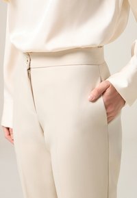 Beige Hochbundhose mit Reißverschluss und Seitentaschen. Glattes Material mit leichtem Glanz und strukturierter Passform.