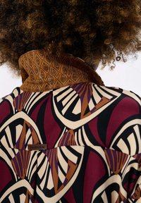 Curly hair with a high collar jacket featuring an abstract geometric pattern in burgundy, cream, black, brown, and purple.