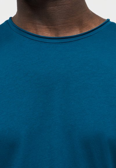 Κοντινό πλάνο του λαιμού και του άνω μέρους του στήθους ενός ατόμου που φοράει μια μπλουζά με στρογγυλή λαιμόκοψη σε τιρκουάζ χρώμα, με απλό φόντο.