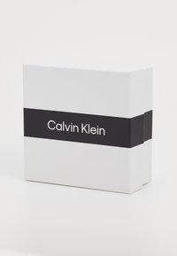 Valkoinen suorakulmainen laatikko matta pinnalla, jossa on musta nauha, johon on painettu "Calvin Klein" -logo myös paksulla, sans-serif -fontilla.
