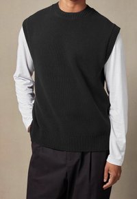 Schwarze gestrickte Weste mit geripptem Kragen und Saum, über einem weißen Langarmshirt getragen, das eine lockere Passform und eine einfarbige Optik zeigt.