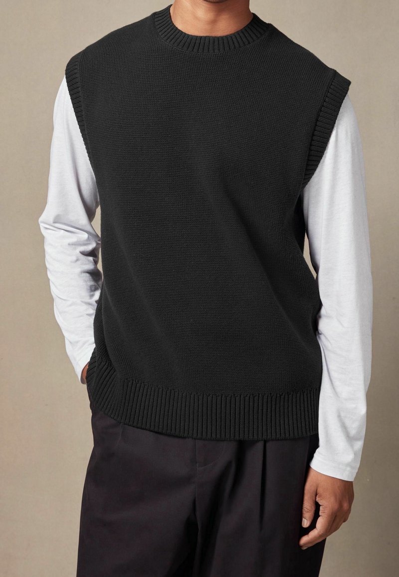 Schwarze gestrickte Weste mit geripptem Kragen und Saum, über einem weißen Langarmshirt getragen, das eine lockere Passform und eine einfarbige Optik zeigt.