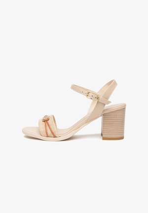 Sandalia beige de punta abierta con tacón ancho, tirante frontal texturizado con nudo decorativo y tirante ajustable en el tobillo con hebilla dorada.