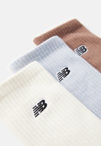 Trois paires de chaussettes côtelées en beige, bleu clair et marron, chacune arborant un logo New Balance noir sur le côté.