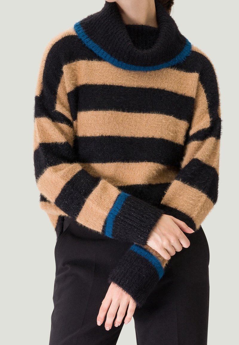 Personne portant un pull à col roulé duveteux avec de larges rayures horizontales beige et noires et des accents bleus au col et aux poignets, associé à un pantalon noir.