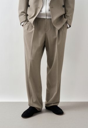 Άνδρας που φοράει μπεζ καρό παντελόνι και σακάκι με τα χέρια στις τσέπες, συνδυασμένο με μαύρα παπούτσια, στέκεται μπροστά σε ένα μονόχρωμο φόντο.