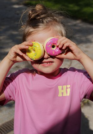 Kind in pinkem Hemd hält gelbe und rosa bestreute Donuts über den Augen im Freien bei Tageslicht.