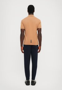 Brun kortermet polo skjorte med krage og sort stripe detalj på baksiden, paired med marine joggebukser og svarte sneakers.