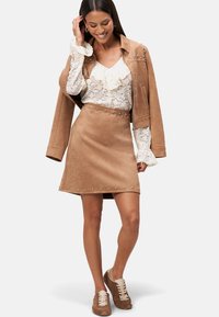 Mini-jupe en suède beige avec des clous argentés le long de l'ourlet, associée à un blouse en dentelle blanche et des baskets brunes. Le mannequin porte une veste assortie.