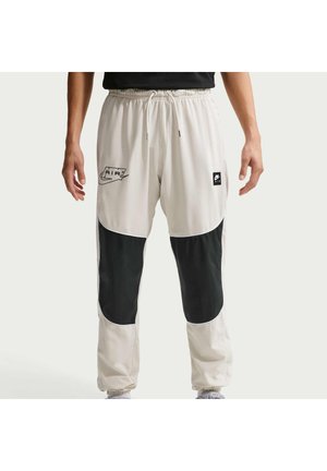 Osoba ubrana w beżowe i czarne spodnie dresowe Nike Air z elastycznym pasem i ściągaczami u dołu, stojąca z ramionami swobodnie opuszczonymi wzdłuż ciała.