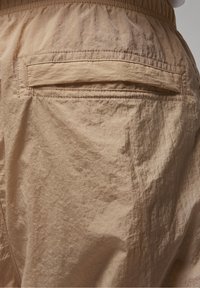 Beige utomhusshorts tillverkade av lätt, skrynklig tyg. Har midjeband i elastisk material och en enda bakficka med ett sydda lock.