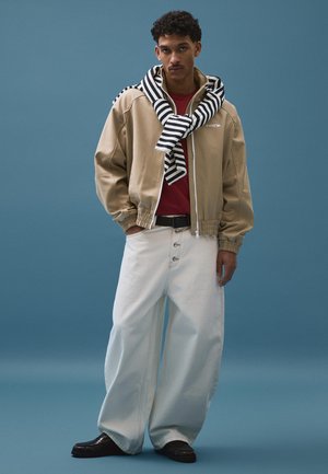 Mladý muž v béžovej bunde, červenej košeli, širokých bielych nohaviciach, čiernych mokasínach a čiernobielom pruhovanom svetri prehodenom cez plecia na modrom pozadí.