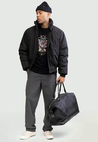 Black puffer jacket over a black graphic t-shirt, gray cargo pants, and beige sneakers. Black leather duffel bag. Knit black beanie.