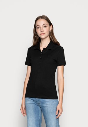 Svart polo-skjorte laget av bomull, med en placket med to knapper, kort erm, og en subtil logo på brystet. Kombinert med lyseblå jeans.