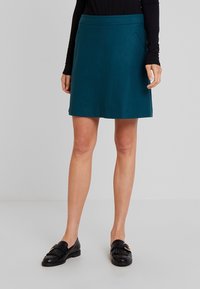 Donna che indossa una gonna teal fino al ginocchio, un top nero a maniche lunghe e mocassini in pelle nera con dettagli in fibbia, in piedi su un pavimento grigio.