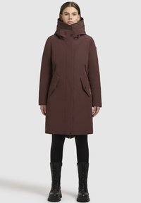 Lang brun frakke med høj krave, lynlås og knaplukning, frontlommer og sort hardware. Båret over sorte leggings og kampstøvler.