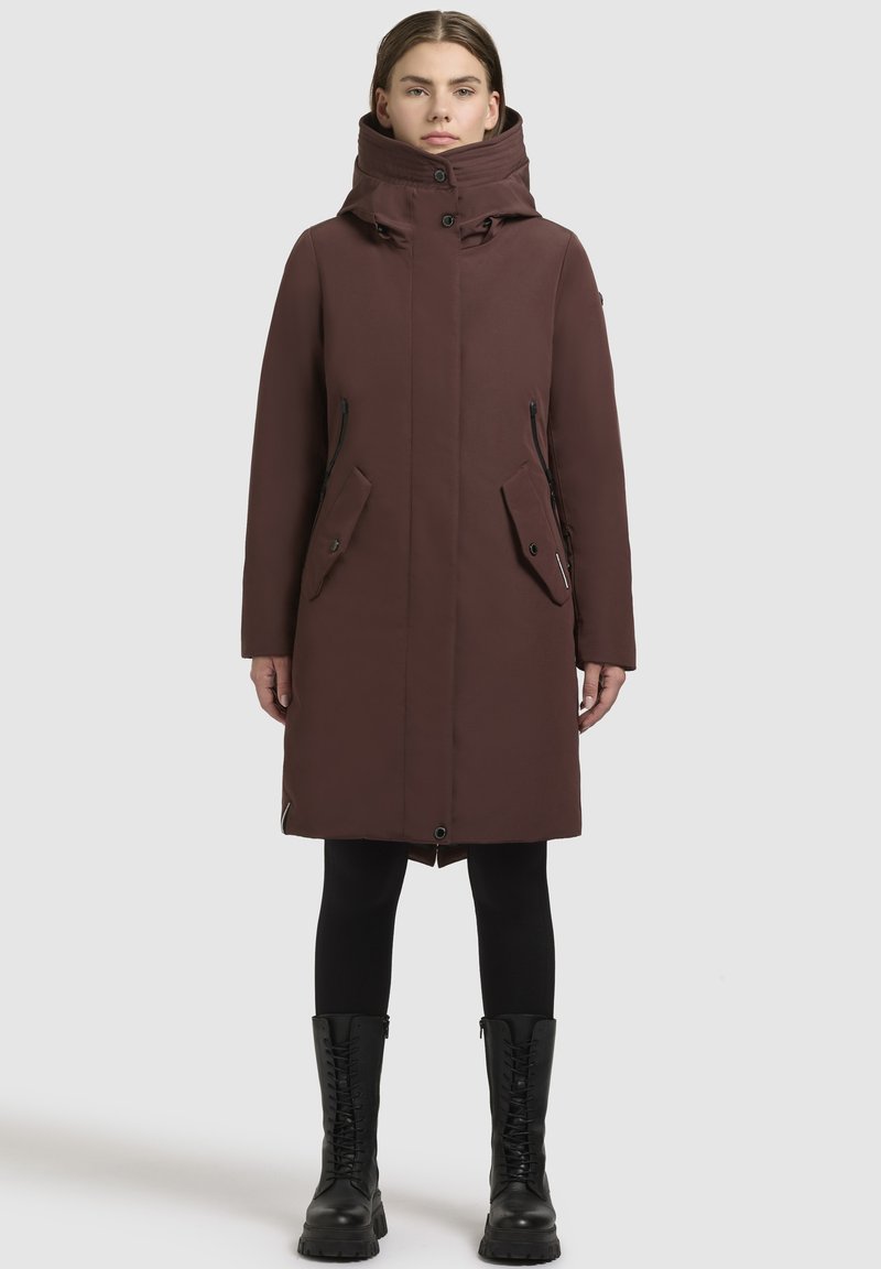 Lungo cappotto marrone con colletto alto, chiusura a zip e bottoni, tasche frontali e dettagli in metallo neri. Indossato sopra leggings neri e stivali militari.