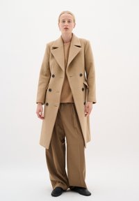 Beige vlněný kabát s dvojitým zapínáním, velkými černými knoflíky, s klopovým límcem, spojený s lehkým svetrem a hnědými kalhotami s širokými nohavicemi.