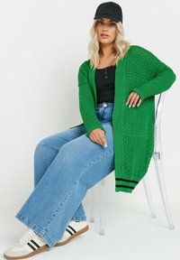 Grön stickad cardigan med öppen front, invecklat diamantmönster, långa ärmar, kombinerad med svart linne och blå vida denimjeans.