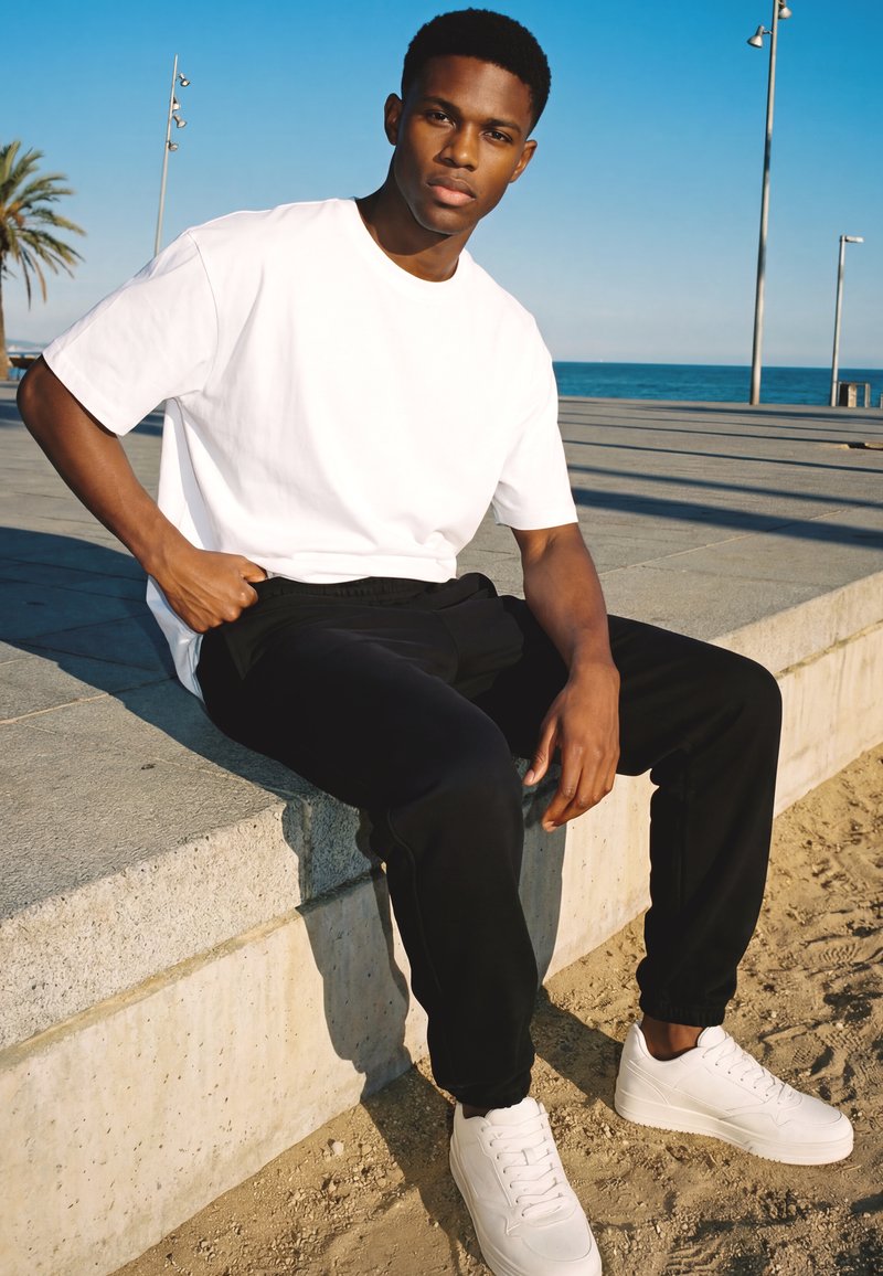 Junger Mann in weißem T-Shirt, schwarzen Hosen und weißen Turnschuhen sitzt auf einer Betonmauer nahe dem Strand mit klarem Himmel im Hintergrund.