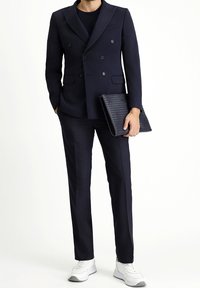 Granatowa marynarka dwurzędowa z klapami typu peak, dopasowane spodnie i teksturowana czarna kopertówka, zestawione z białymi butami sportowymi.