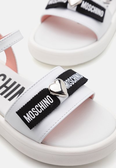 Sandalias blancas con una correa elástica negra grabada con "MOSCHINO", un acento en forma de corazón plateado y un forro interior rosa. Diseño de suela plana.
