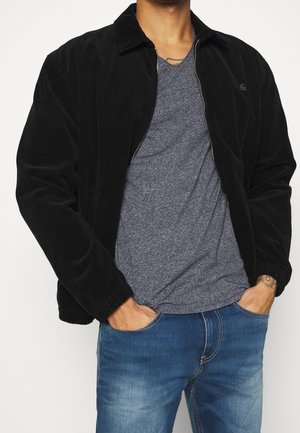 Black corduroy jacket with a collar and front zipper, paired with a gray crew-neck t-shirt. Worn over blue jeans, showcasing a relaxed fit.