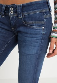 Mörkblå denim skinny jeans med hög midja, featuring två knäppen, framfickor och kontrasterande sömmar. Slät textur.