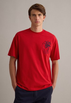 Giovane uomo con indosso una maglietta rossa con un motivo floreale blu scuro sul petto, in piedi con le mani nelle tasche dei pantaloni blu navy.