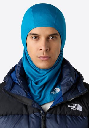 Teal balaklava gjord av stretchmaterial, åtsittande design, bärs med en mörk puffjacka som har logotyper och dragkedjor på bröstområdet.